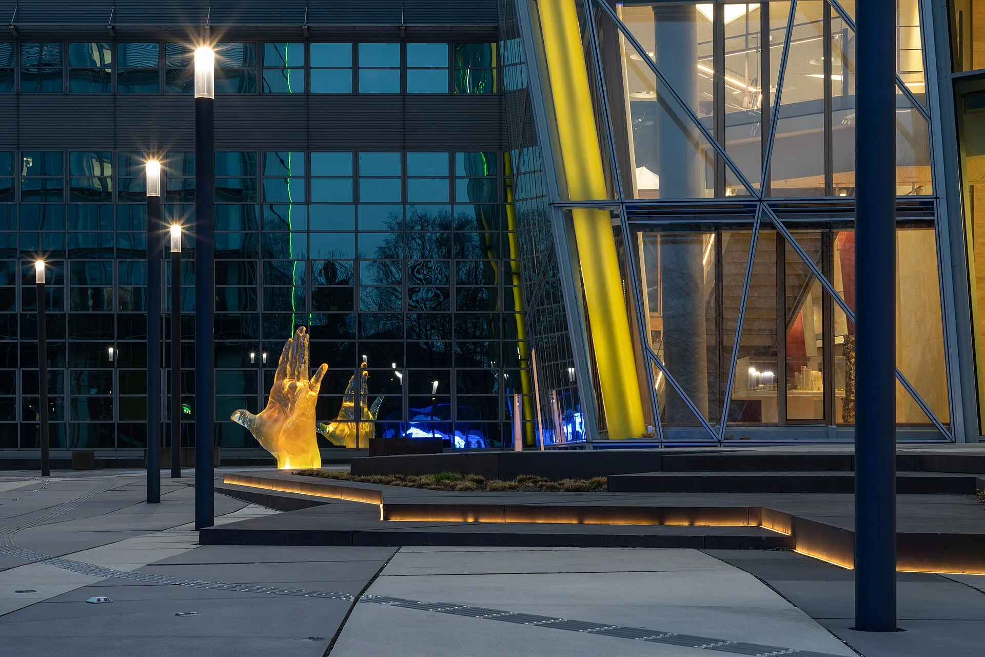 Office forecourt at dusk with glowing step edges, slim light columns and a lit glass facade